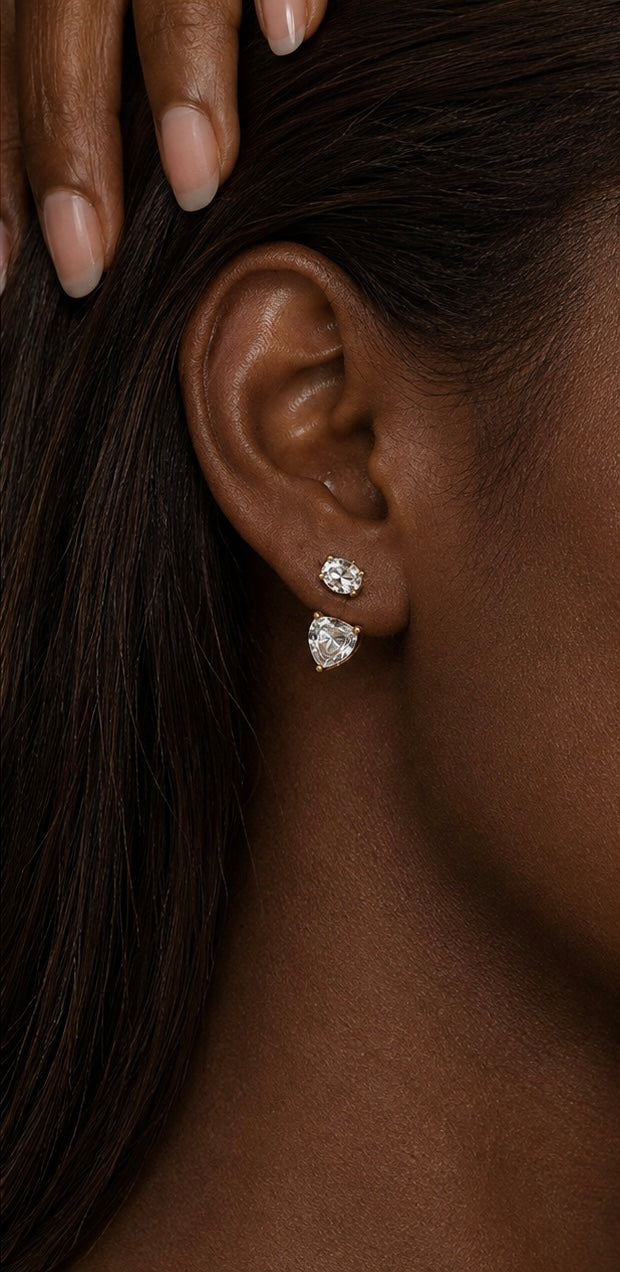 Close-up of a model's ear wearing a pair of small diamond stud earrings with a pear-cut diamond drop, set in yellow gold.